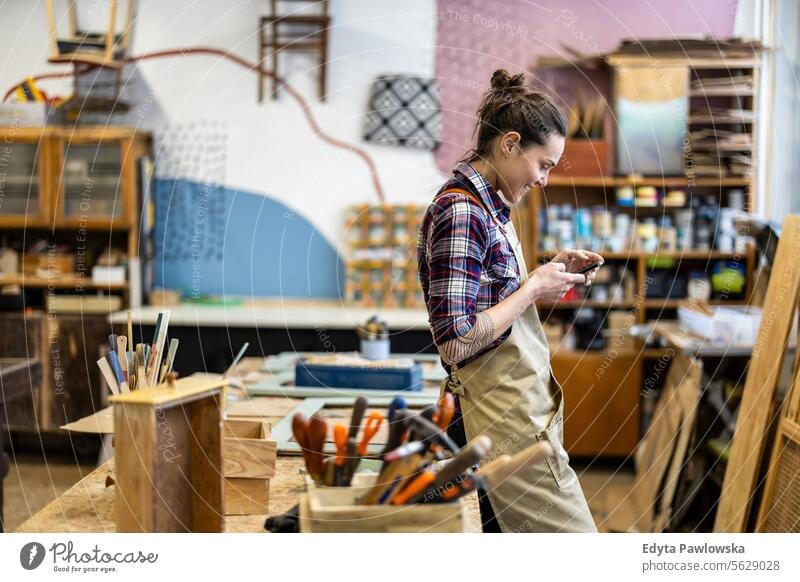 Porträt einer jungen Schreinerin, die in ihrer Werkstatt ein Mobiltelefon benutzt Möbel Zimmerer wiederherstellend Schreinerei Holz Stuhl Gebäude Herstellung