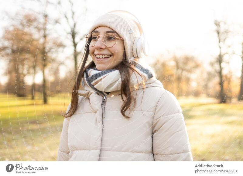 Lächelnde Frau mit Kopfhörern, die im Herbst im Park Musik hört zuhören Gesang positiv genießen Freizeit warme Kleidung Brille Drahtlos jung Glück Hut