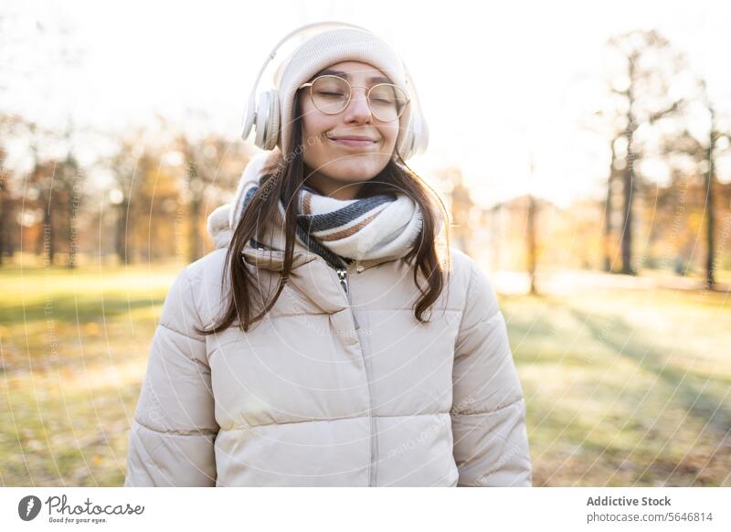 Lächelnde Frau mit Kopfhörern, die im Herbst im Park Musik hört zuhören Gesang positiv genießen Freizeit warme Kleidung Brille Drahtlos jung Glück Hut