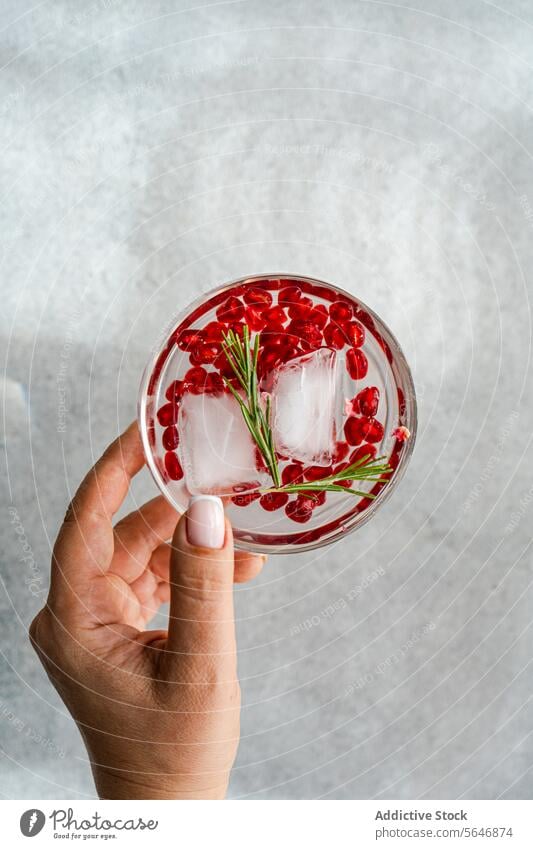 Anonymes Bild einer Hand, die einen Gin Tonic hält, in einem konkreten Hintergrund Cocktail Granatapfel Samen Rosmarin Glas Alkohol trinken Eis grau texturiert