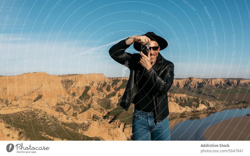 Fotograf in Lederjacke rahmt Blick mit Retro-Kamera ein Mann Glück Hut Rahmen Hände Ansicht Lächeln malerisch analog Schlucht Fotokamera Blauer Himmel stylisch