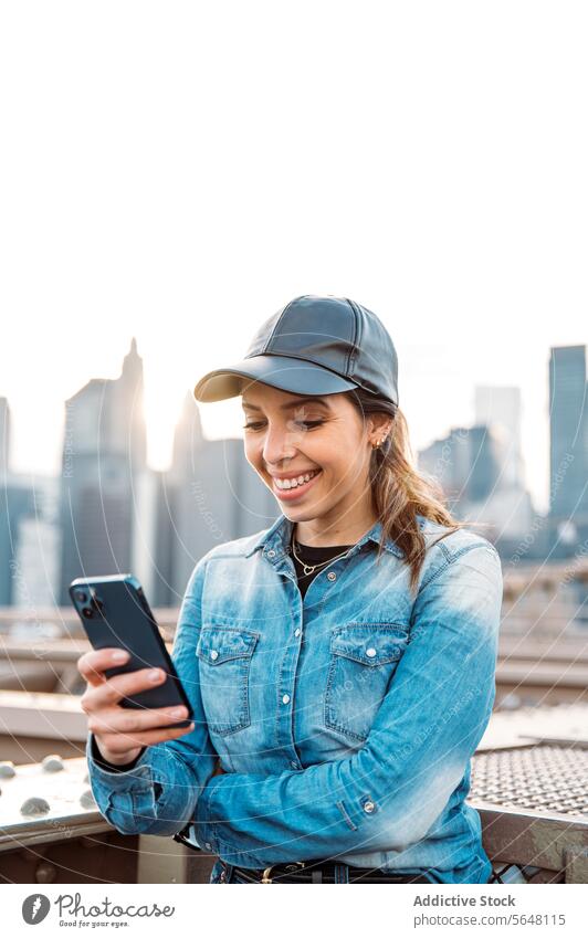 Frau mit Smartphone vor Manhattan-Kulisse Sonnenbrille Verschlussdeckel Lächeln Skyline Stadtbild New York State im Freien urban Brücke Mode Freizeit lässig