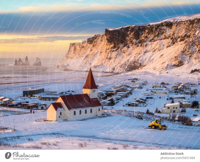 Der Sonnenuntergang wirft ein warmes Licht über ein kleines isländisches Dorf mit einer Kirche zwischen schneebedeckten Klippen und dem Meer Schnee MEER Island