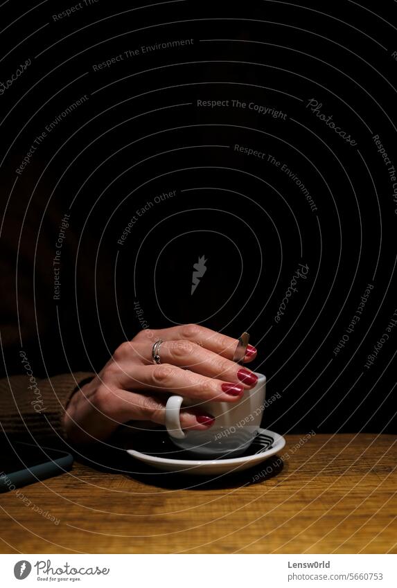 Die Hand einer Frau, die aus der Dunkelheit heraus eine Tasse Kaffee hält schwarz Kaffeetasse Finger Fingernägel Nagellack Nägel Ringe Rampenlicht Heißgetränk