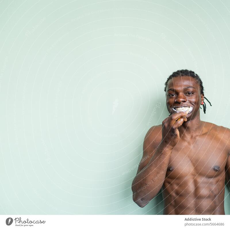 Ein schwarzer Mann ohne Hemd mit Nasenring und Ohrringen putzt sich die Zähne vor einem hellgrünen Hintergrund Zahnpflege Zahnbürste Hygiene Zahncreme