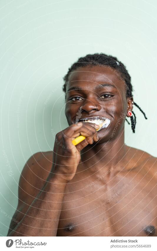 Porträt hemdloser schwarzer Mann mit Nasenring und Ohrringen putzt sich die Zähne vor einem hellgrünen Hintergrund Zahnpflege Zahnbürste Hygiene Zahncreme