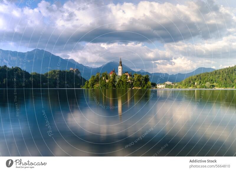 Im ruhigen Wasser des Bleder Sees spiegeln sich an einem bewölkten Tag die idyllische Kircheninsel und die Burg inmitten der Julischen Alpen Gelassenheit