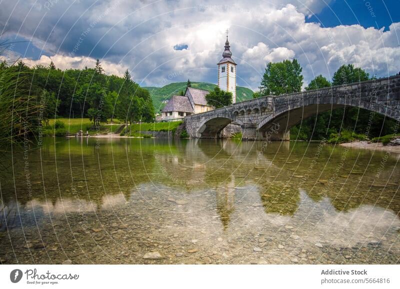 Eine Steinbrücke spannt sich über das kristallklare Wasser des Bohinjer Sees mit der Kirche St. Johannes der Täufer und den Bergen im Hintergrund Brücke