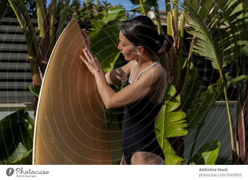 Seitenansicht einer fokussierten Surferin, die ein Surfbrett berührt, während sie in einem Garten mit tropischen Blättern steht Frau Vorbereitung berührend