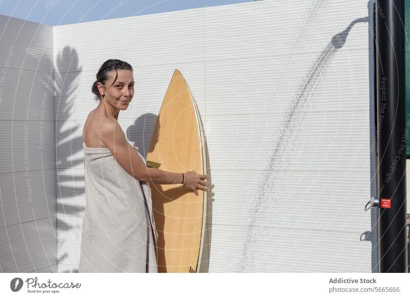 Lächelnde Frau im Handtuch, die nach dem Duschen neben einem Surfbrett steht und in die Kamera schaut im Freien Sommer Freizeit Erholung Hinterhof Badeanzug