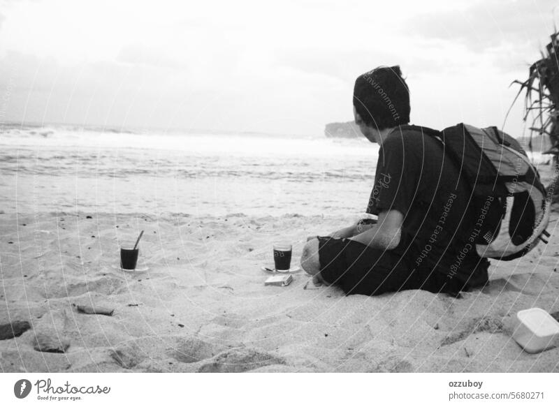 Junger erwachsener Mann sitzt allein auf Sand und schaut weit weg auf Seite am Meer Strand mit einem Glas Tasse Kaffee und Daypack Tasche auf dem Rücken Person