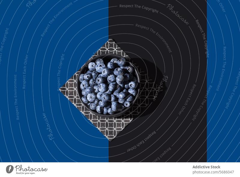 Draufsicht auf eine Schale mit saftigen Blaubeeren auf einer geometrisch gemusterten Serviette vor einem kontrastierenden blauen und schwarzen diagonalen Hintergrund