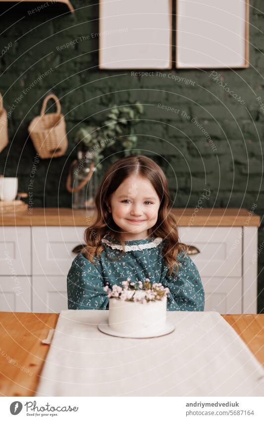 Adorable kleines Kind Mädchen trägt festliche Kleid nimmt einen Bissen aus einer dekorierten Blumen Namen Kuchen auf einer Geburtstagsfeier. Glücklich lächelnd Kind leckt weiße Creme aus ihrem schmutzigen Gesicht und zeigt Daumen nach oben drinnen