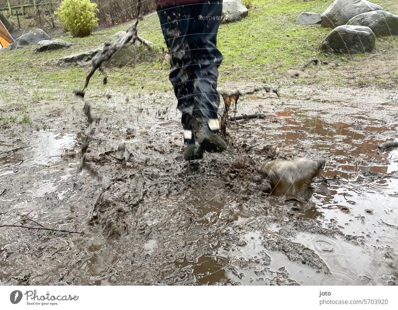 Kind mit Regenhose springt in einer matschigen Pfütze regnerisch regnerisches Wetter Herbst herbstlich herbstliche Stimmung achterbahnfahrt grau Regentag