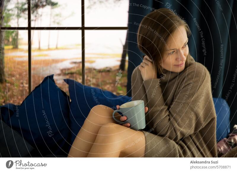 Hübsche junge Frau liest ein Buch und trinkt einen Morgenkaffee zu Hause, während sie am Fenster sitzt, hinter dem sich eine Herbstlandschaft befindet
