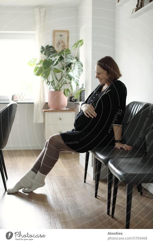 seitliches Portrait einer jungen, schwangeren, lachenden Frau, die auf einem Stuhl im Wohnzimmer sitzt junge Frau Schwangerschaft schwangere Frau sitzen Freude