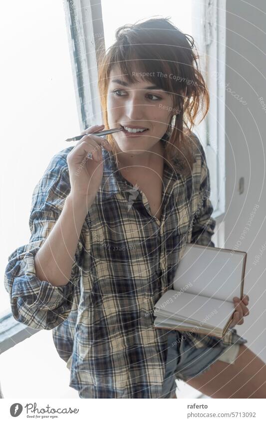 Eine nachdenkliche junge Frau hält einen Stift und ein leeres Notizbuch in der Hand, bereit, kreative Ideen zu notieren. Bleistift Notebook kariertes Hemd