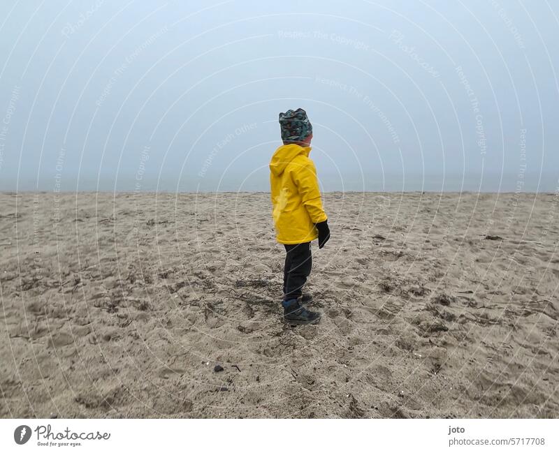 Kind steht am Strand mit Blick auf das Meer an einem nebeligen Tag Herbst herbstlich herbstliche Stimmung trüb Außenaufnahme leer Natur Wasser trist Oktober