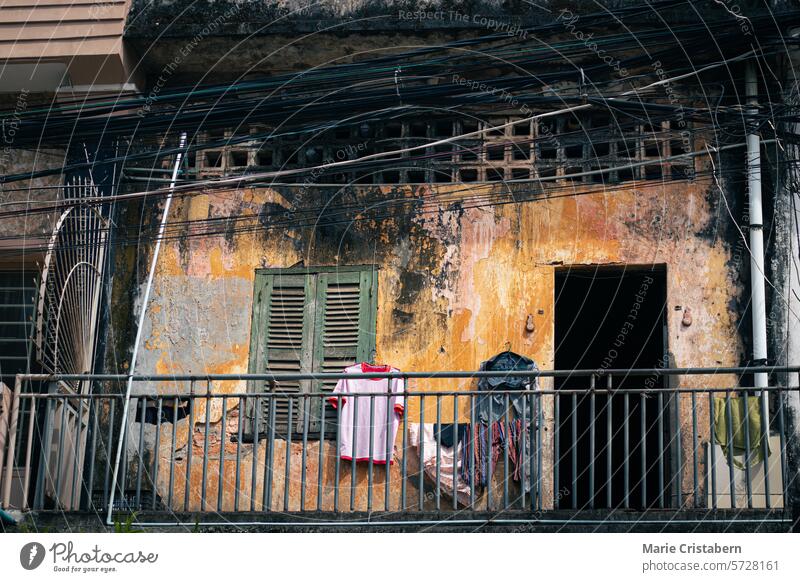 Verwitterter Balkon eines alten rustikalen Kolonialgebäudes mit aufgehängter Wäsche, grünen Fensterläden, Geländer und sichtbaren Drähten über dem Kopf, die eine offene Szene aus dem täglichen Leben der Khmer in Kampot, Kambodscha, zeigen
