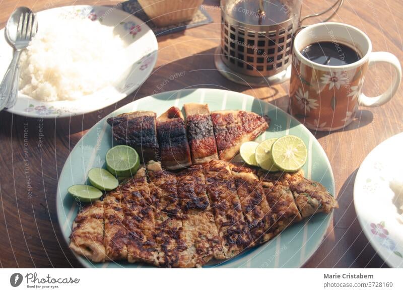 Ein morgendliches Frühstück mit gegrilltem Fisch, Reis und Kaffee in einem Garten, mit einer französischen Presse auf einem Holztisch unter der Sommersonne