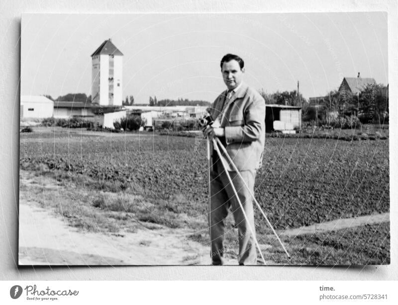 Mann mit Stativ und Kamera an einem Feldweg Landschaft Acker Horizont Stadtrand Turm Architektur Jacke Blick in die Kamera Porträt