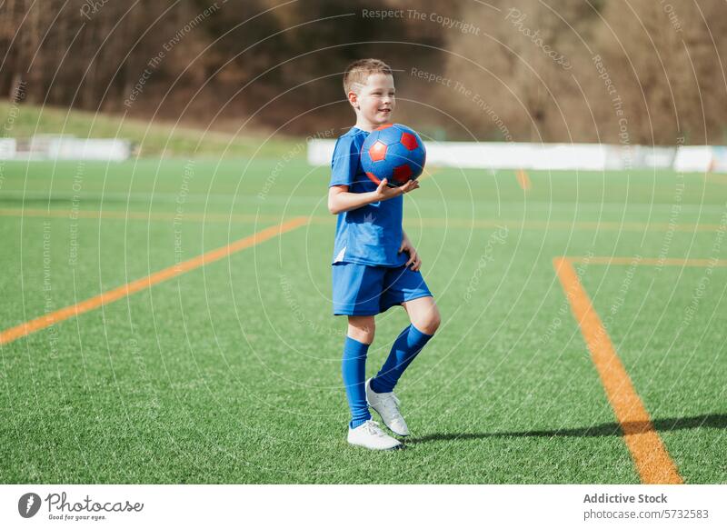 Junger Fußballspieler hält einen Ball auf dem Spielfeld Feld Sport jung Spieler Uniform blau grün Gras Stehen Selbstvertrauen Kind im Freien Team Freizeit