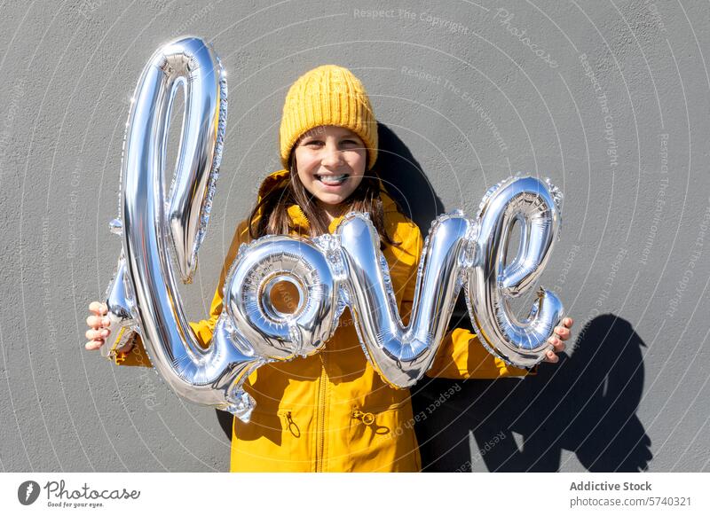 Lächelnde Frau hält silberne Liebesballon-Buchstaben Silber Luftballon gelb Hut Mantel grau Wand glänzend Glück Feier positiv Freude Emotion Spaß festlich