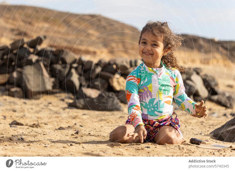 Ein glückliches Kleinkind Mädchen in einem bunten Badeanzug spielt mit Sand am Strand, mit Felsen und Hügeln im Hintergrund spielen farbenfroh Glück im Freien