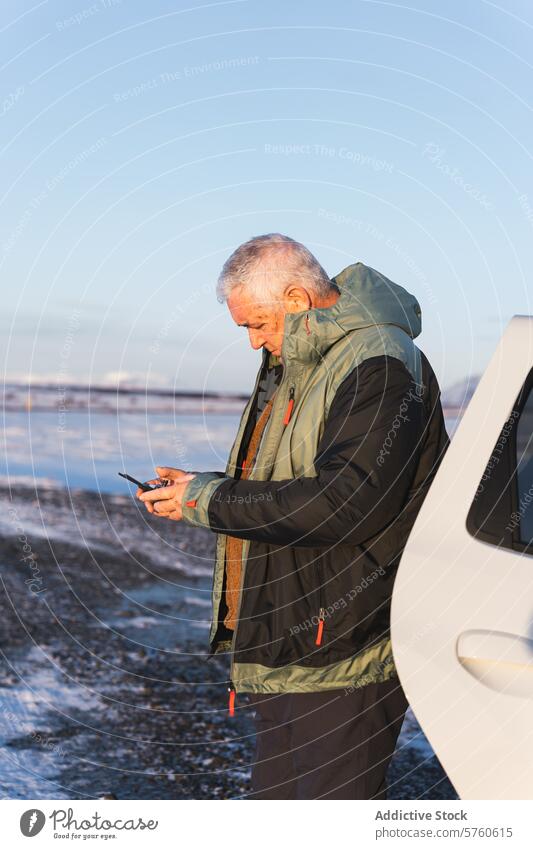 Ein älterer Reisender beschäftigt sich mit seinem Smartphone, während er im weichen Licht der isländischen Wintersonne neben einem Fahrzeug auf eisigem Terrain steht