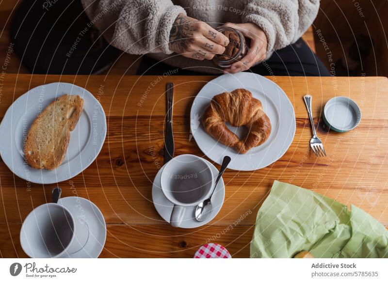 Gemütlicher Frühstückstisch mit frischem Gebäck und tätowierten Händen Tisch Croissant Kaffee Tasse Teller Tattoo Hand Holz Brot Gabel Messer gemütlich Morgen