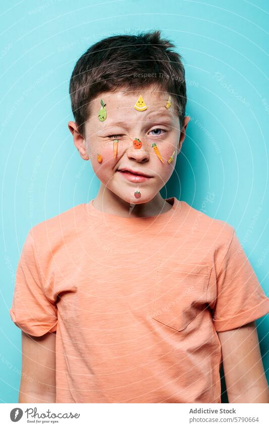 Junge mit verspielten Fruchtaufklebern im Gesicht spielerisch Glück türkisfarbener Hintergrund posierend jung niedlich farbenfroh Klebstoff Gesichtsausdruck