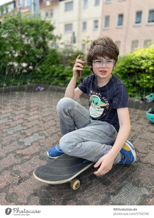 Junge sitzt auf einem Skateboard und telefoniert mit dem Handy Kind Kindheit natürlich authentisch spass Außenaufnahme Zukunft frech Blick urban Stadt Mensch