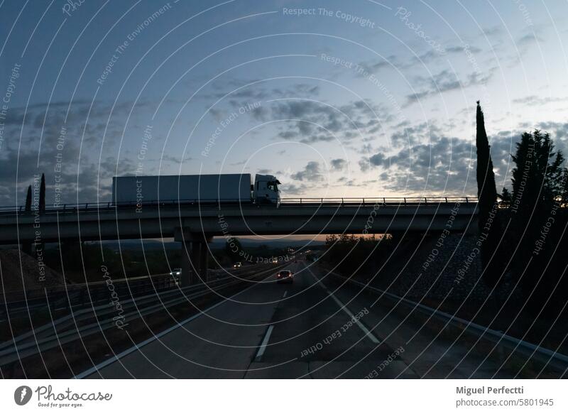 Autobahn in der Abenddämmerung mit Fahrzeugen und einer Brücke, auf der ein Lkw fährt, Seitenansicht des Lkw. Lastwagen Verkehr logistisch Sonnenuntergang