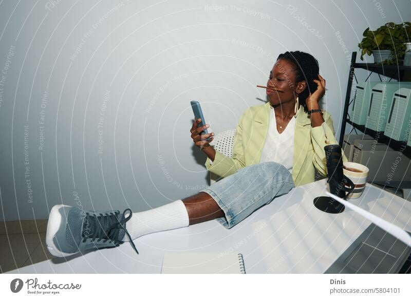 Businesswoman sitting with foot on office desk and taking selfie holding pencil between lips and nose fun photograph funny posing work playful businesswoman