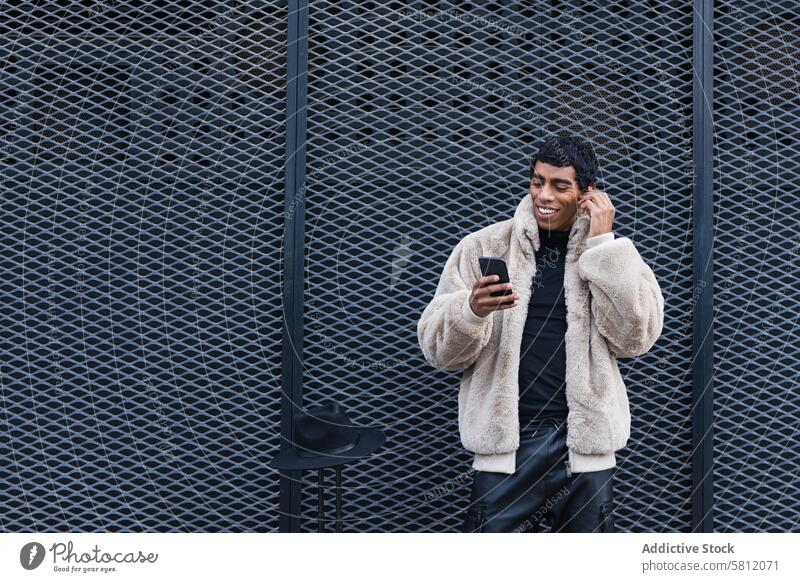 Inhalt stilvoller ethnischer Mann mit Smartphone im Ohrhörer auf der Straße Ohrstöpsel zuhören Gesang genießen trendy Drahtlos benutzend Apparatur Gerät