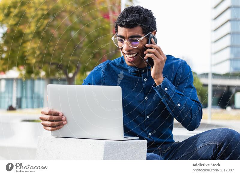 Glücklicher ethnischer Geschäftsmann Freiberufler, der an einem Laptop arbeitet und mit einem Mobiltelefon in der Stadt spricht Mitarbeiter Mann freiberuflich
