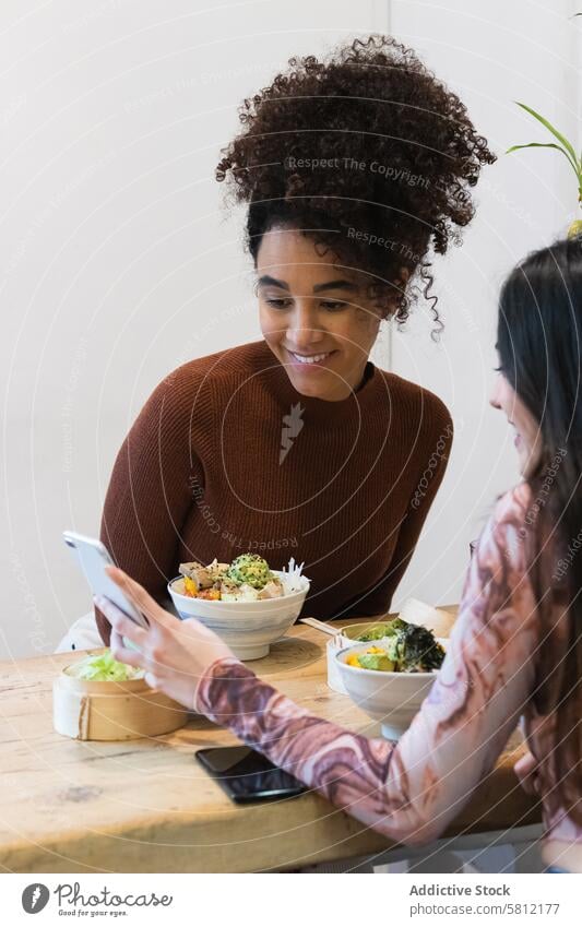 Lächelnde multiethnische Frauen beim Essen im Restaurant Zusammensein Freund Smartphone teilen Zeit verbringen Freundschaft benutzend Lebensmittel vielfältig