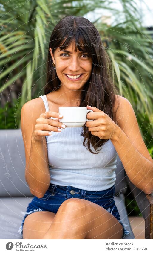 Eine Frau schaut in die Kamera, während sie auf einer Couch sitzt und eine weiße Tasse hält. Sie lächelt und genießt ihren Kaffee modern Erwachsener attraktiv