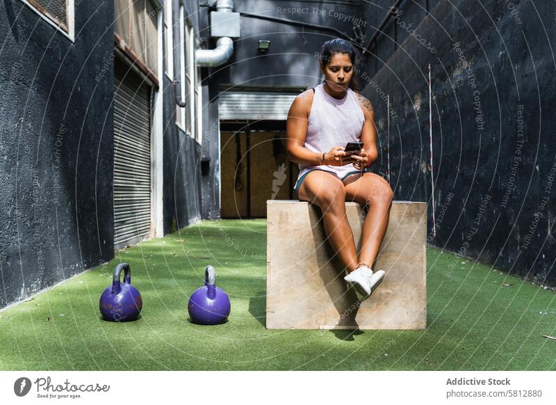 Ethnische Frau, die auf einem Sprungkasten auf ihrem Smartphone surft benutzend Fitness Training Kettlebell Pause Browsen ernst Fitnessstudio hölzern