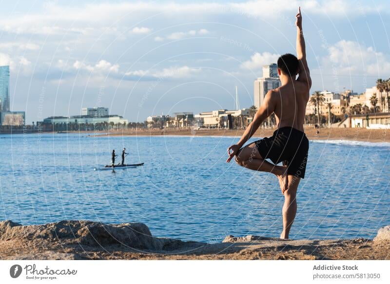 Mann ohne Hemd balanciert in Yoga-Pose am Strand Baumhaltung vriksasana üben Asana Gleichgewicht stehen männlich Mudra gestikulieren Lifestyle Wellness