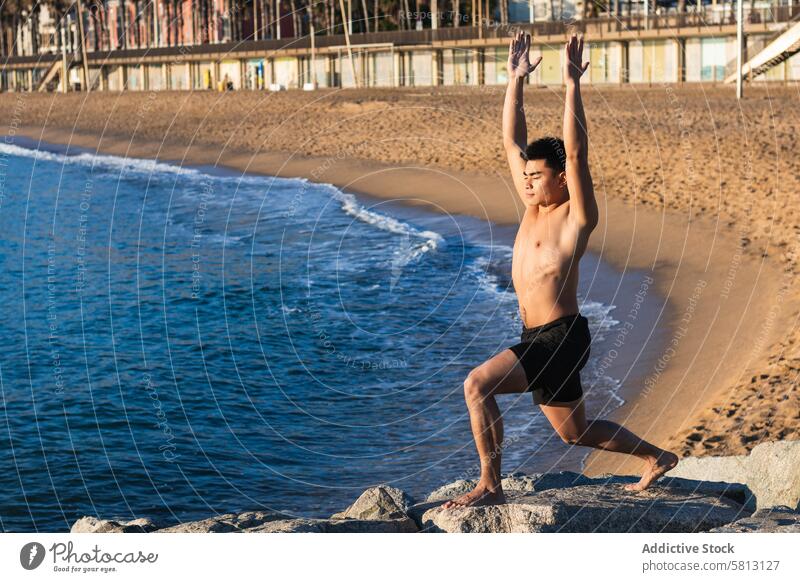 Ethnischer Mann in Yogapose am Meeresufer Strand Krieger-Pose virabhadrasana üben Asana Gleichgewicht stehen männlich asiatisch ethnisch ohne Hemd jung
