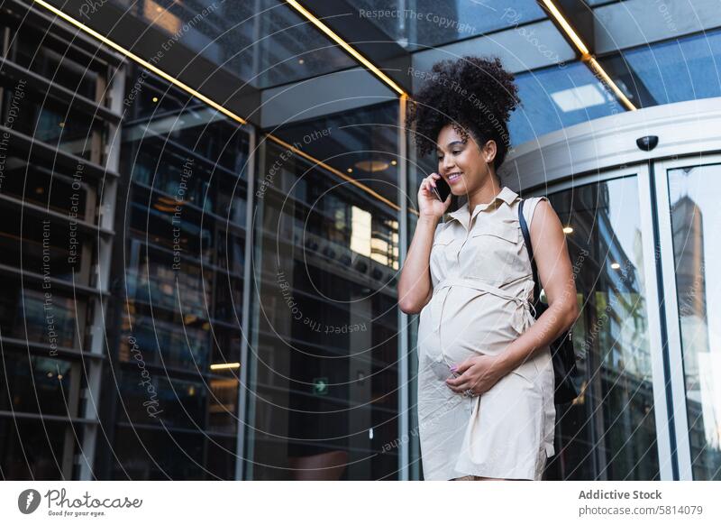 Lächelnde schwarze schwangere Frau, die mit ihrem Smartphone spricht benutzend reden Gespräch positiv Bauch Talkrunde warten sprechen Funktelefon Gerät