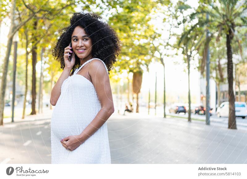 Lächelnde schwarze schwangere Frau, die mit ihrem Smartphone spricht benutzend reden Gespräch positiv Bauch Talkrunde warten sprechen Funktelefon Gerät