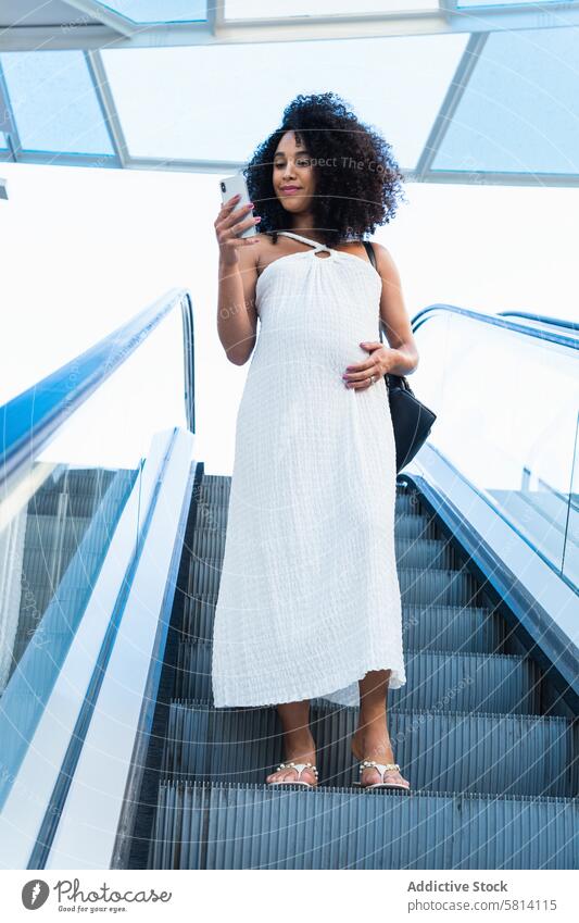 Schwangere schwarze Frau durchsucht Smartphone auf Rolltreppe schwanger benutzend Bauch Browsen Anschluss vorwegnehmen online warten Schwangerschaft erwarten