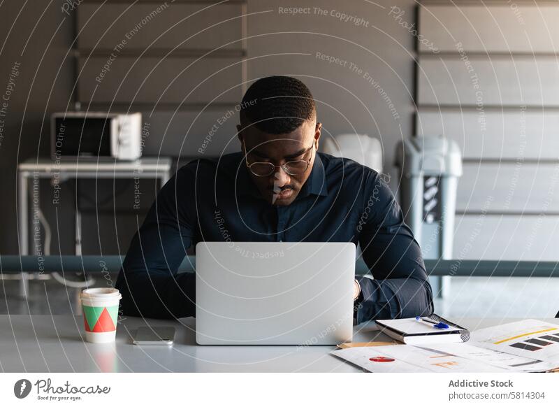 Schwarzer Mann beim Videochat auf Tablet in der Nähe des Laptops am Arbeitsplatz Tablette Browsen Netbook Tisch Büro Schriftstück Pappbecher Zeitgenosse schwarz