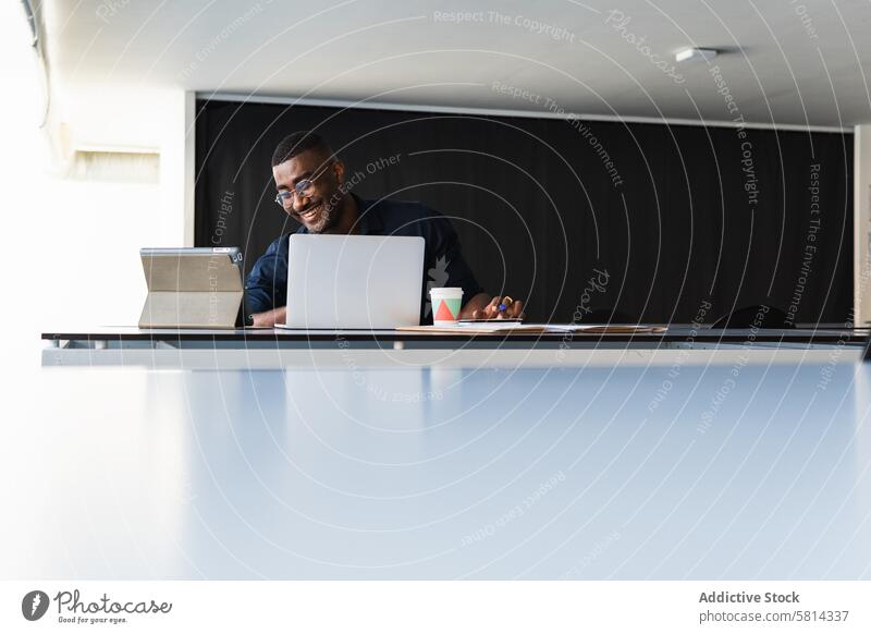 Schwarzer Mann beim Videochat auf Tablet in der Nähe des Laptops am Arbeitsplatz Tablette Videoanruf Netbook Tisch Glück Büro Schriftstück Pappbecher