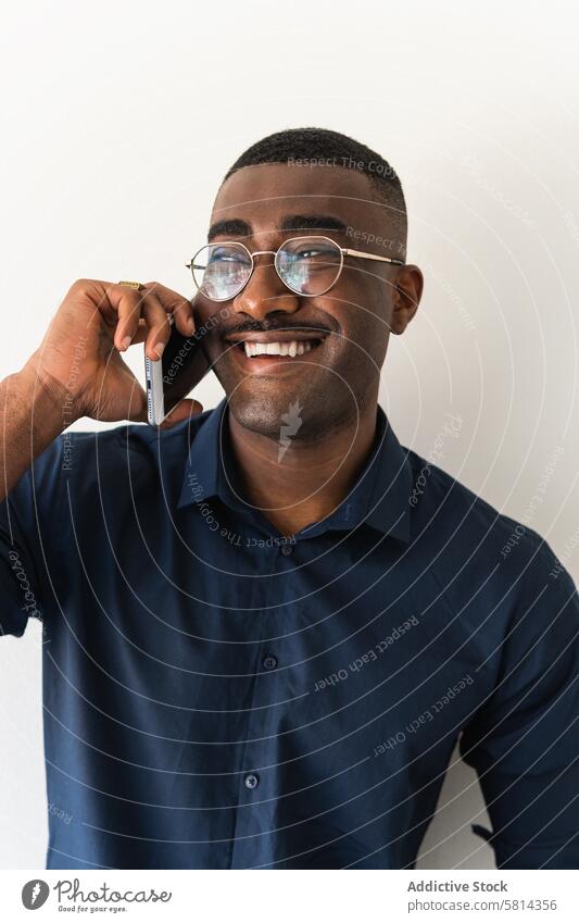 Schwarzer Mann mit Telefon in der Nähe der weißen Wand Smartphone gut gekleidet modern Lächeln Glück froh Brille Studioaufnahme Netbook schwarz Afroamerikaner