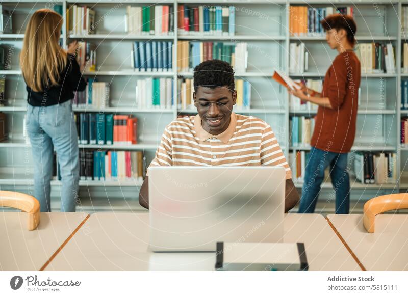 Schwarzer Student mit Laptop in der Bibliothek Schüler benutzend Hausaufgabe Anweisung Lächeln lernen Klassenkamerad Bücherschrank pflücken Buch Teenager
