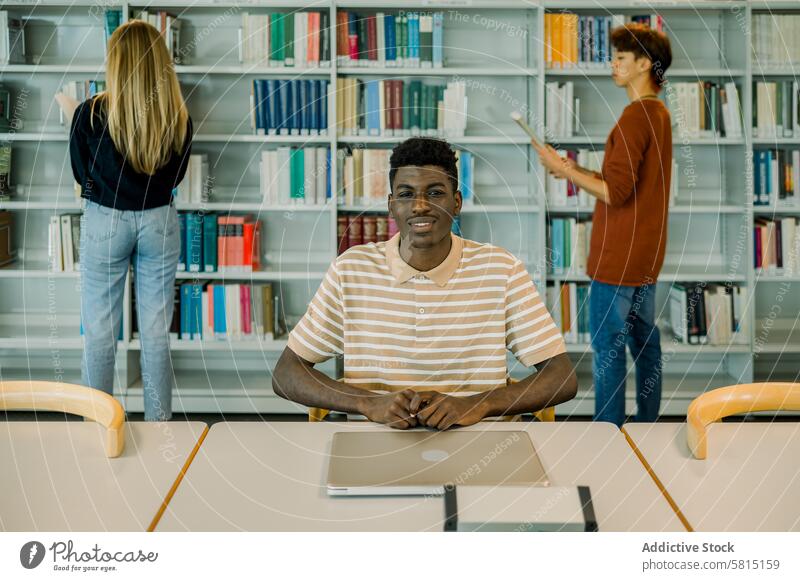 Glücklicher junger schwarzer Mann sitzt mit geschlossenem Laptop Schüler Bibliothek Lächeln positiv Bücherregal Bücherschrank Bildung lernen Universität