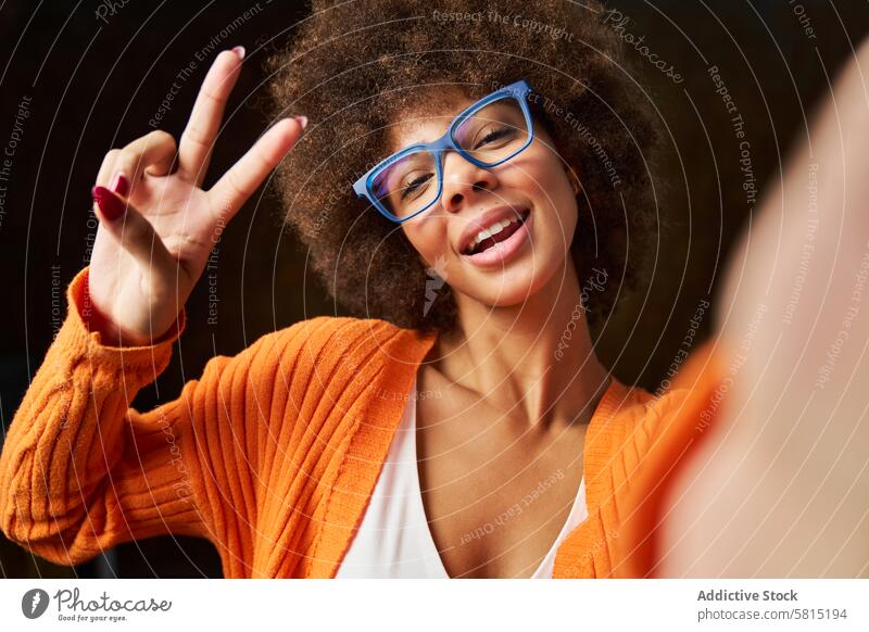 eine junge Frau mit Afro-Haar, die ein Selbstporträt mit Brille und einer Siegesgeste aufnimmt Afro-Look Behaarung Glück gestikulieren dunkel Hintergrund unter